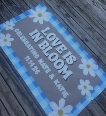 Hand-painted banner lying on a wooden deck reading "Love is in bloom, celebrating Nate and Katie, 11.14.26", with white daisies on a blue gingham border.