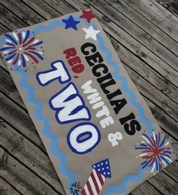 Hand-painted kraft paper banner lying on a wooden deck reading "Cecilia is red, white and two", with painted fireworks, stars, and a firecracker.