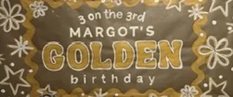 Hand-painted banner reading "3 on the 3rd, Margot's golden birthday" in gold and white lettering, with white stars and flowers.